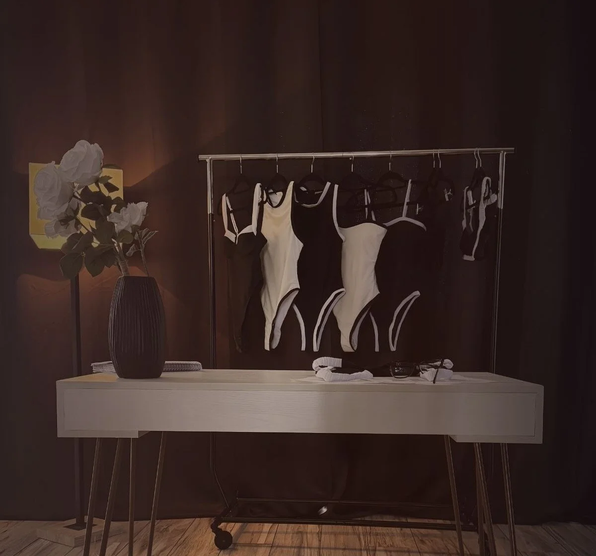 A clothing rack containing black and white bodysuits, a vase with a white flower on a white table, and sunglasses, with a dark wall in the background.