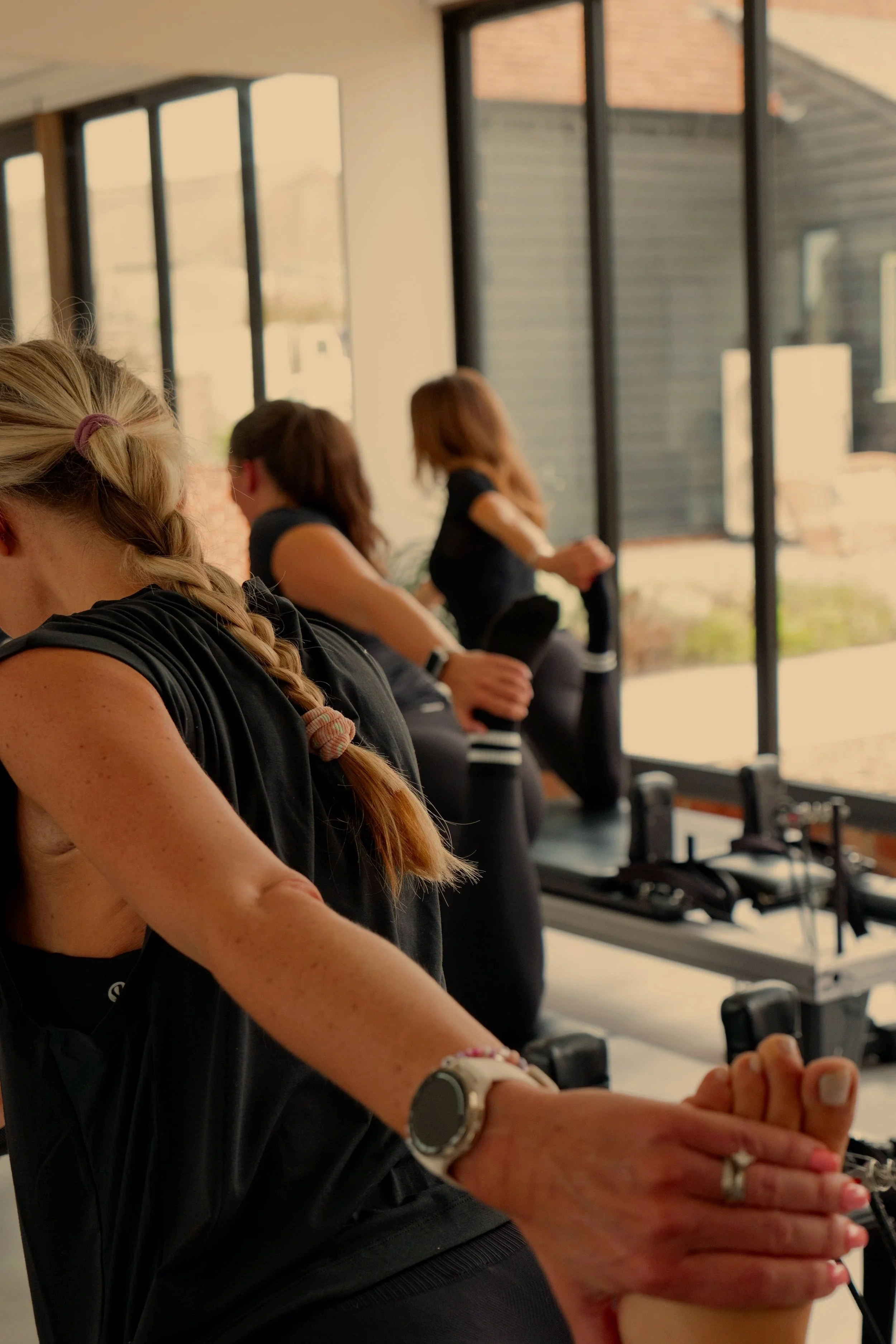 Group of women participating in a fitness class, doing stretching exercises while seated on a bench near large windows.