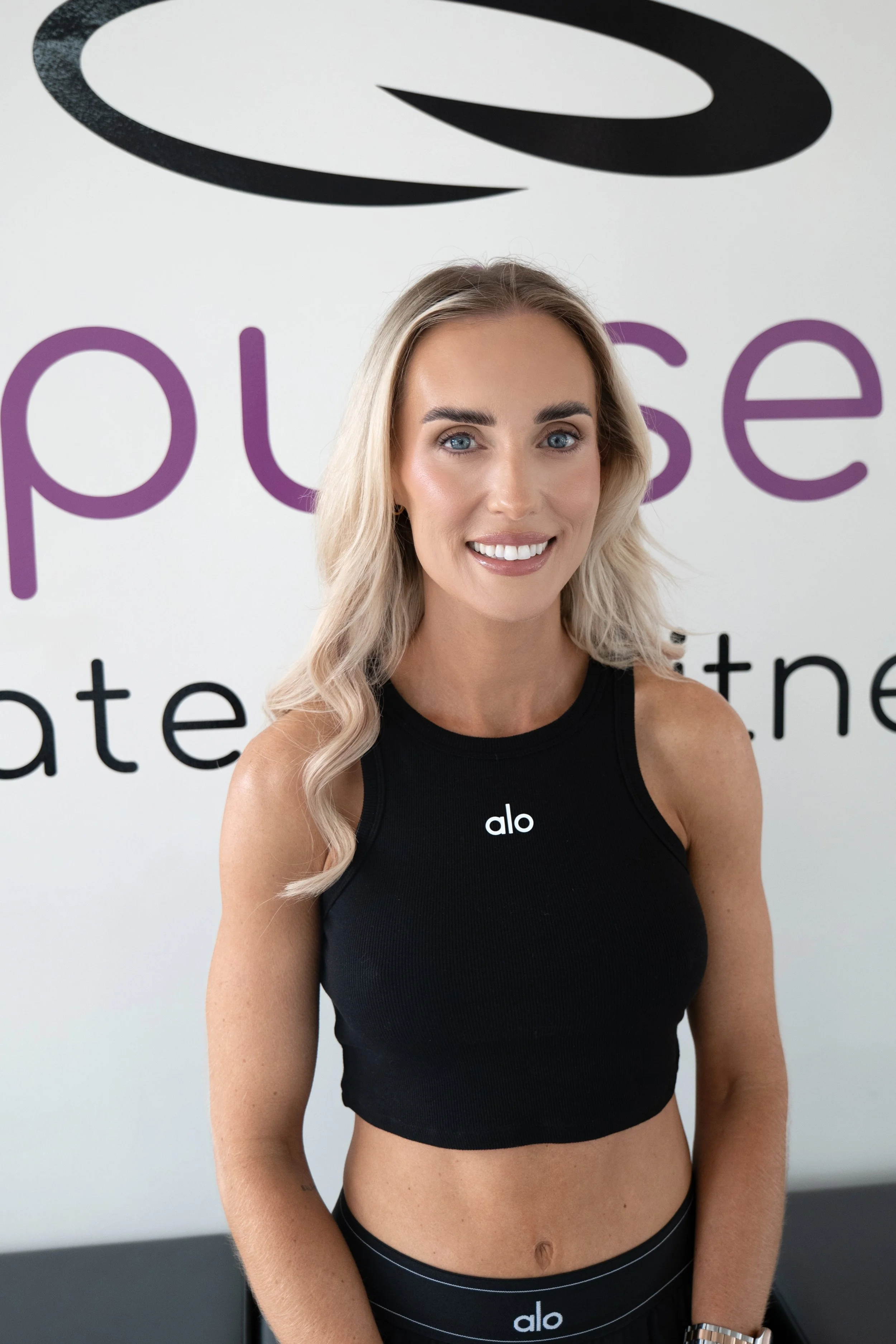 A young woman with blonde wavy hair and blue eyes, smiling, standing indoors in front of a wall with the logo and text for 'Alo' and 'pulse'. She is wearing a black athletic crop top with the 'alo' logo on the chest and matching 'alo' branded black leggings.