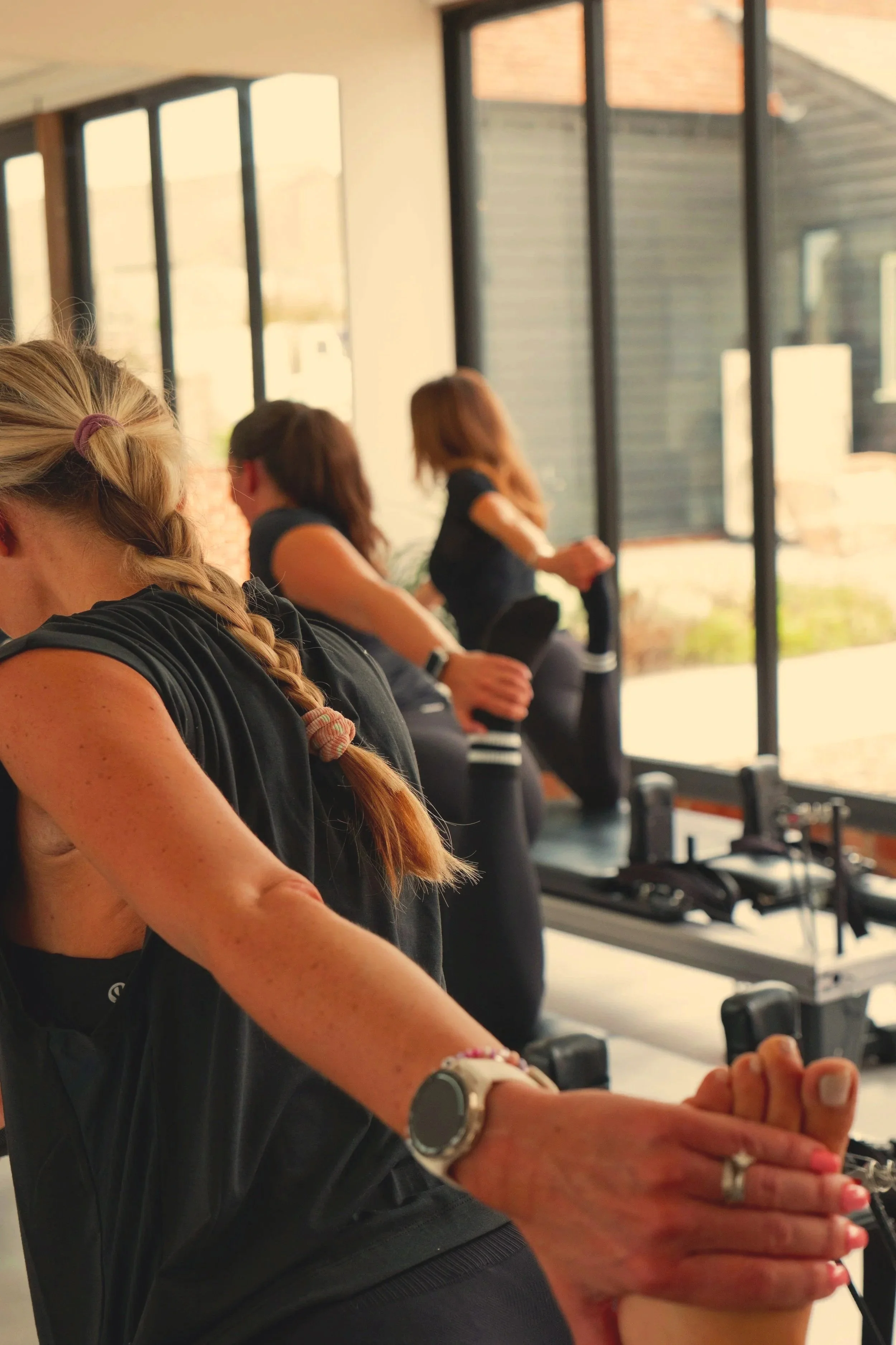 A group of women exercising together in a fitness class, stretching their legs while seated on exercise machines near large windows.