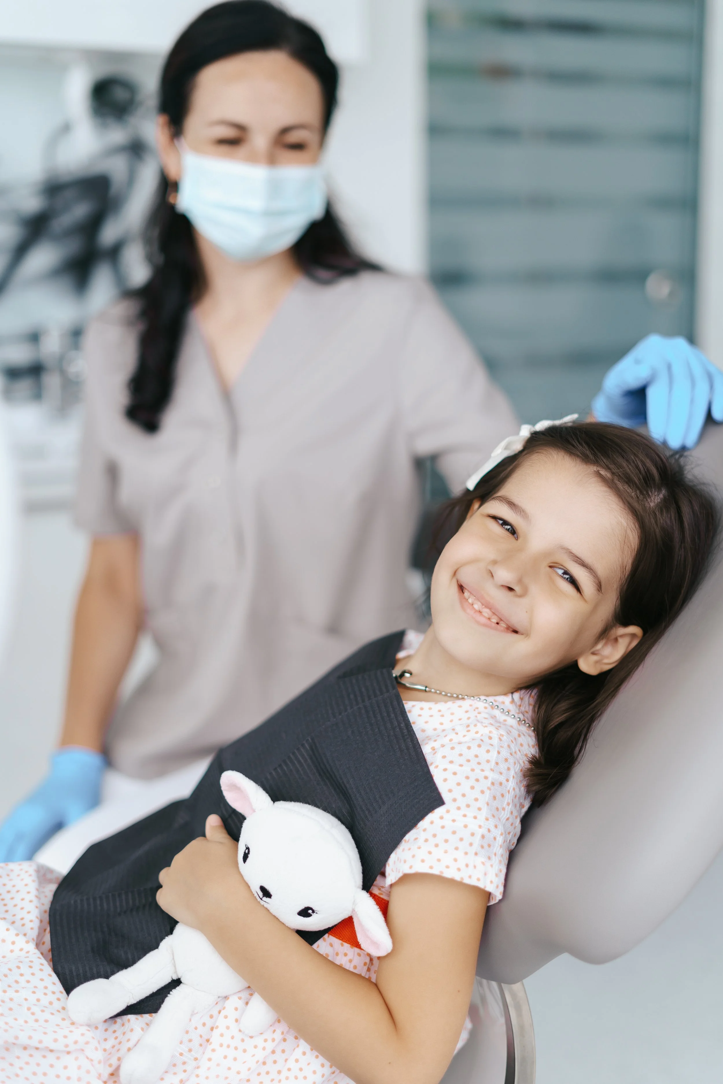 A young girl is lying in a dental chair, smiling and holding a stuffed white bunny; a dental professional wearing a face mask and gloves is standing next to her, preparing or examining her.