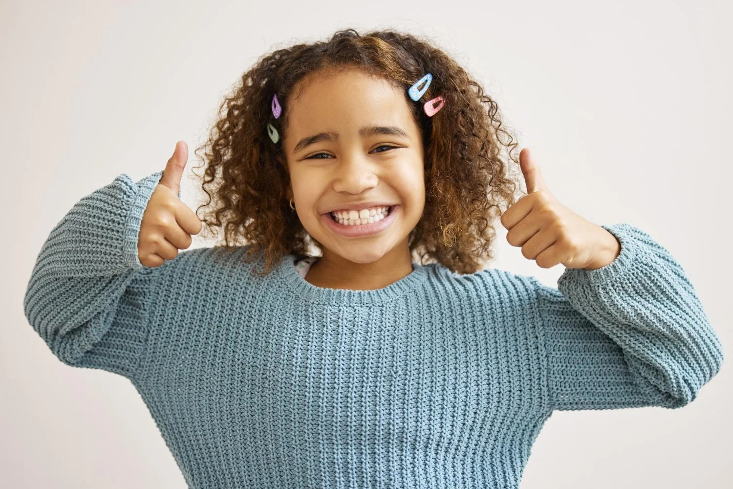 A young girl with curly hair wearing a blue sweater giving a thumbs-up and smiling at the camera.