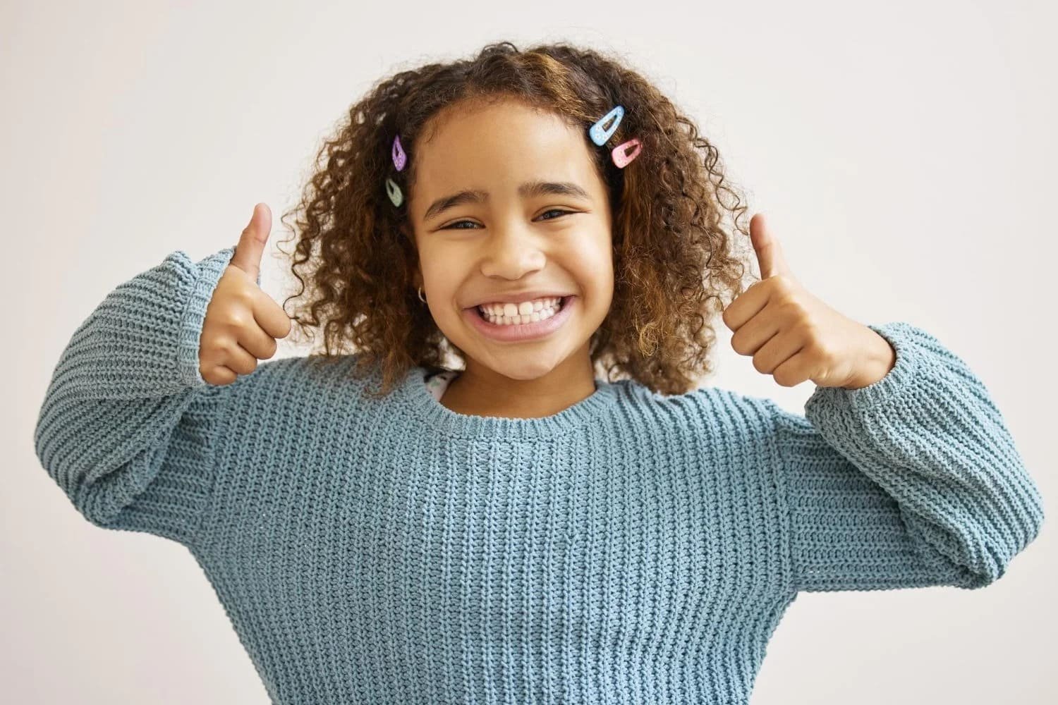 A young girl with curly hair, wearing a light blue sweater and colorful hair clips, smiling and giving thumbs up.