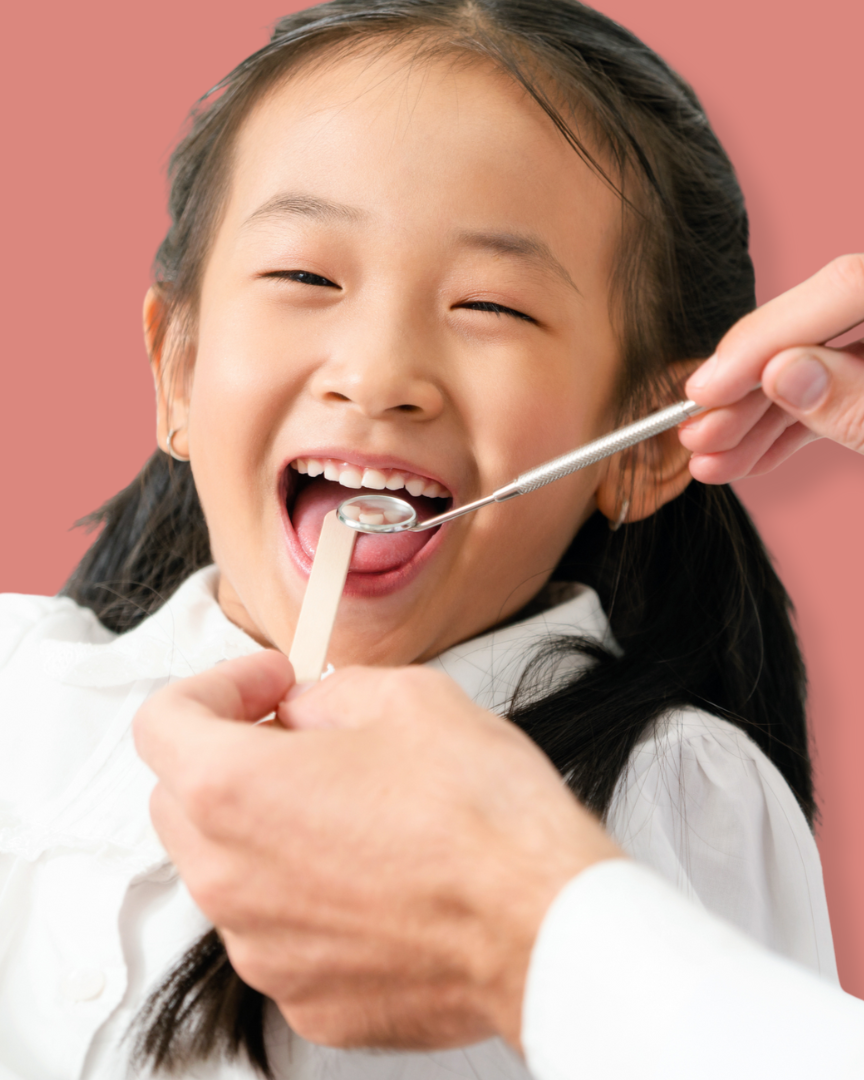 A young girl with black hair smiling during a dental checkup, with a dental mirror and probe near her open mouth against a pink background.