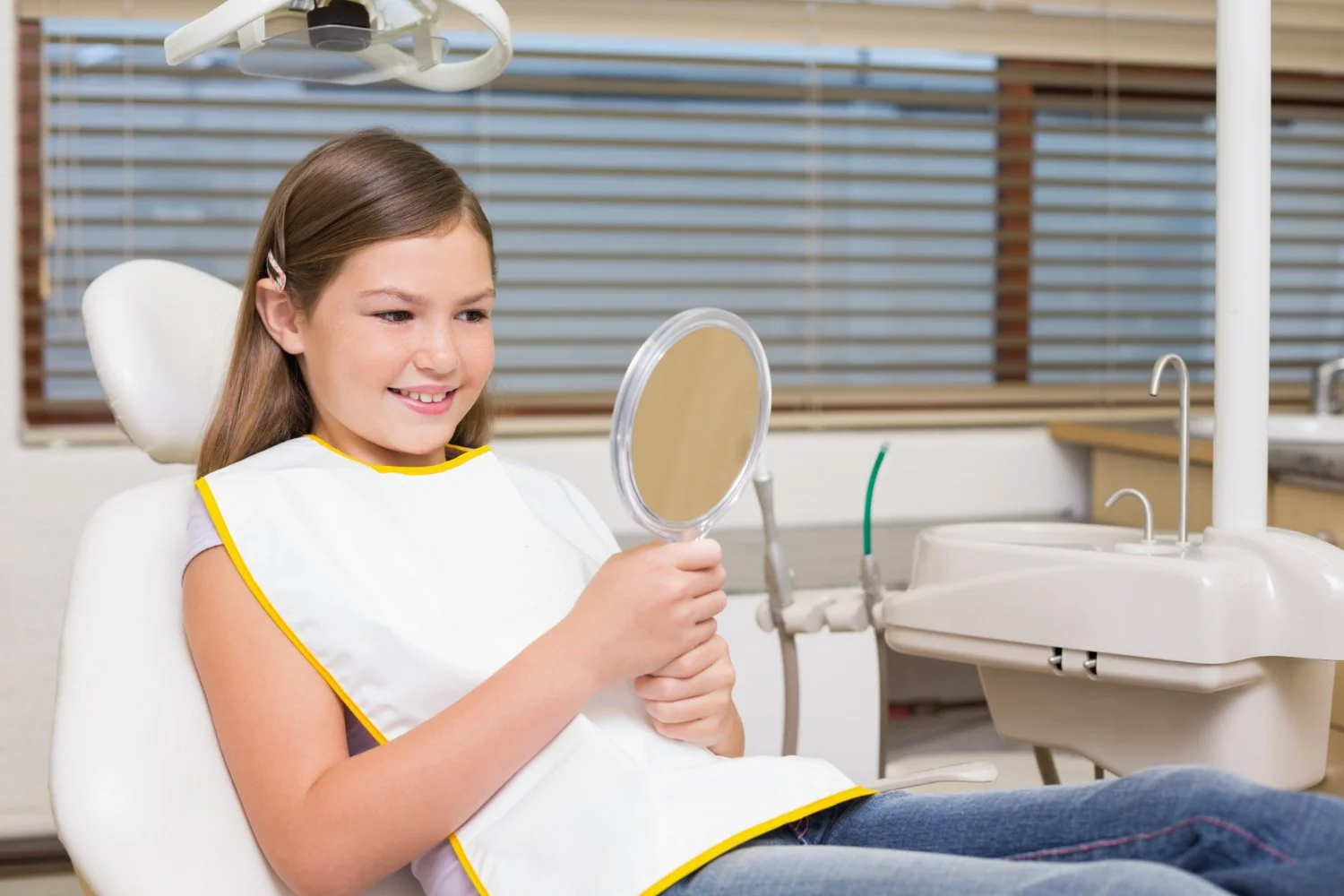 A young girl with brown hair, wearing a white dental bib with yellow trim, is sitting in a dentist's chair holding a mirror and smiling at her reflection. The dental clinic background includes dental equipment, a sink, and wooden blinds covering a window.