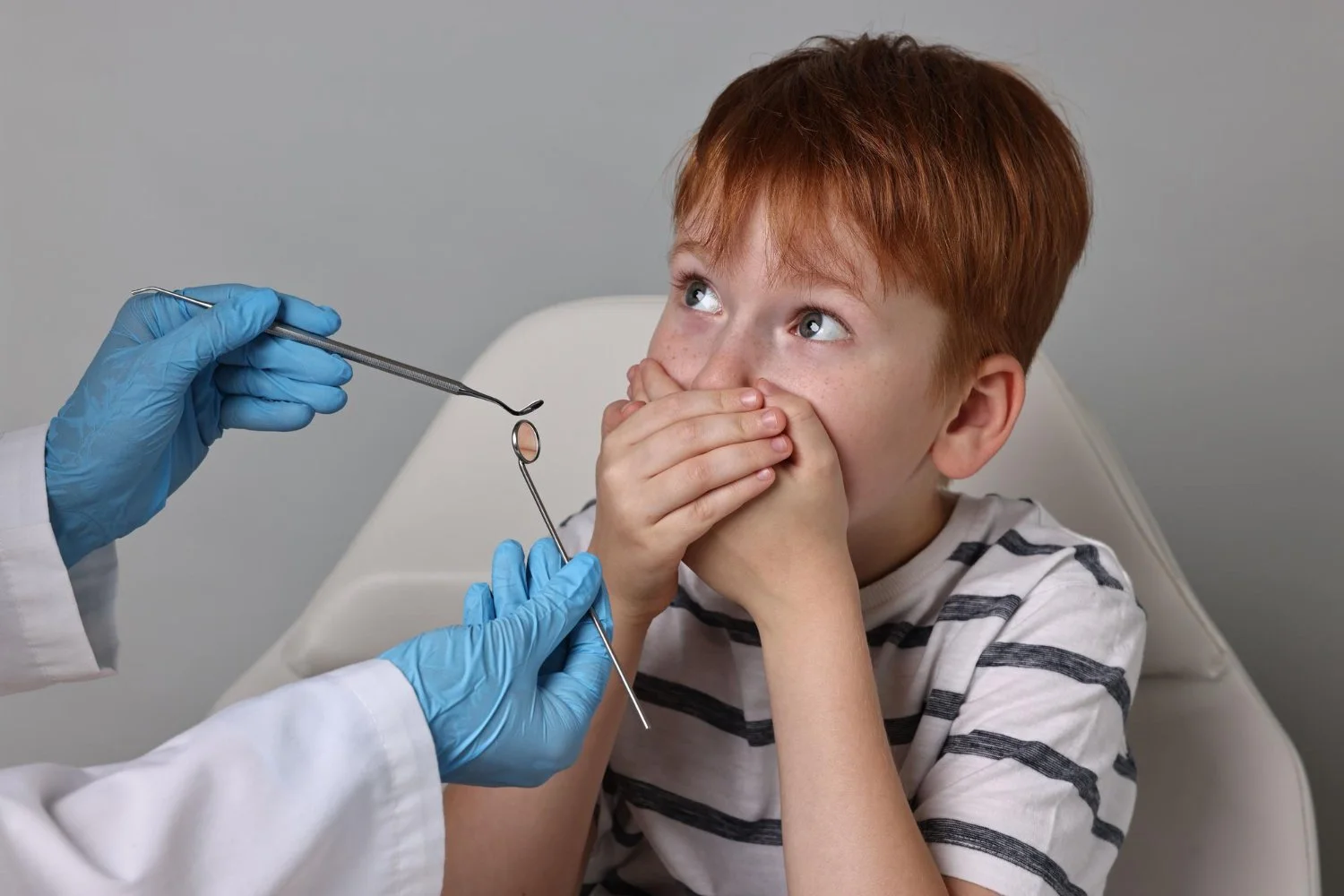 A young boy with red hair and freckles sitting in a chair, covering his mouth with his hands, as a healthcare professional wearing blue gloves and a white coat prepares to hold his nose with a medical tool for a nasal exam.