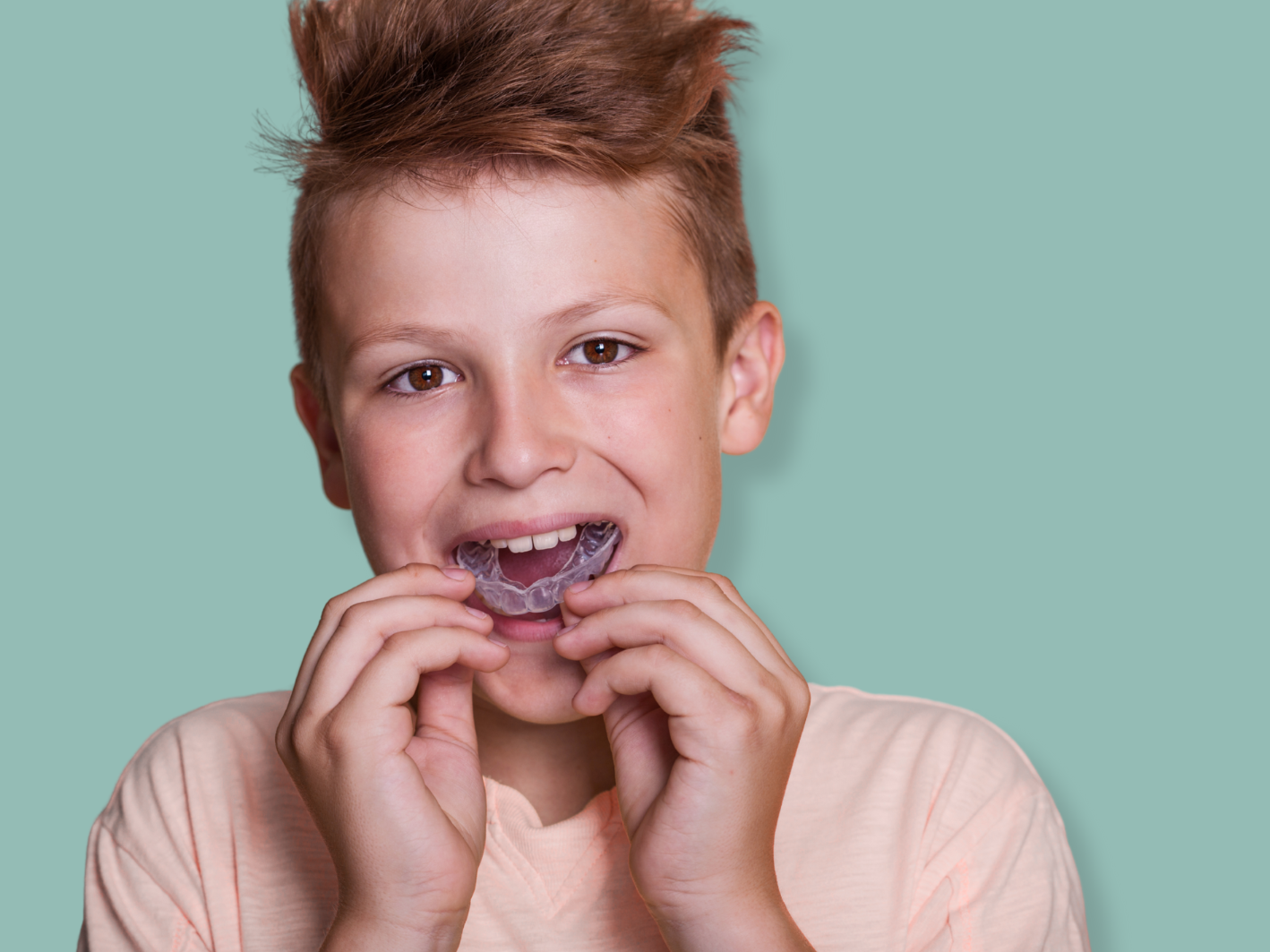 A young boy with short, light brown hair holding a clear dental night guard in his mouth, smiling, against a light turquoise background.
