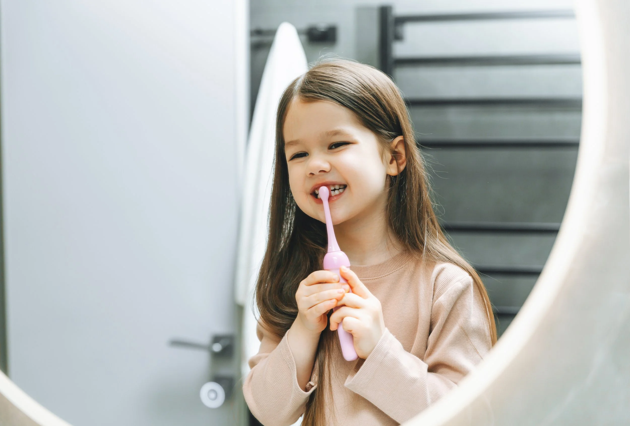 Young girl brushing her teeth with a pink toothbrush in front of a mirror.