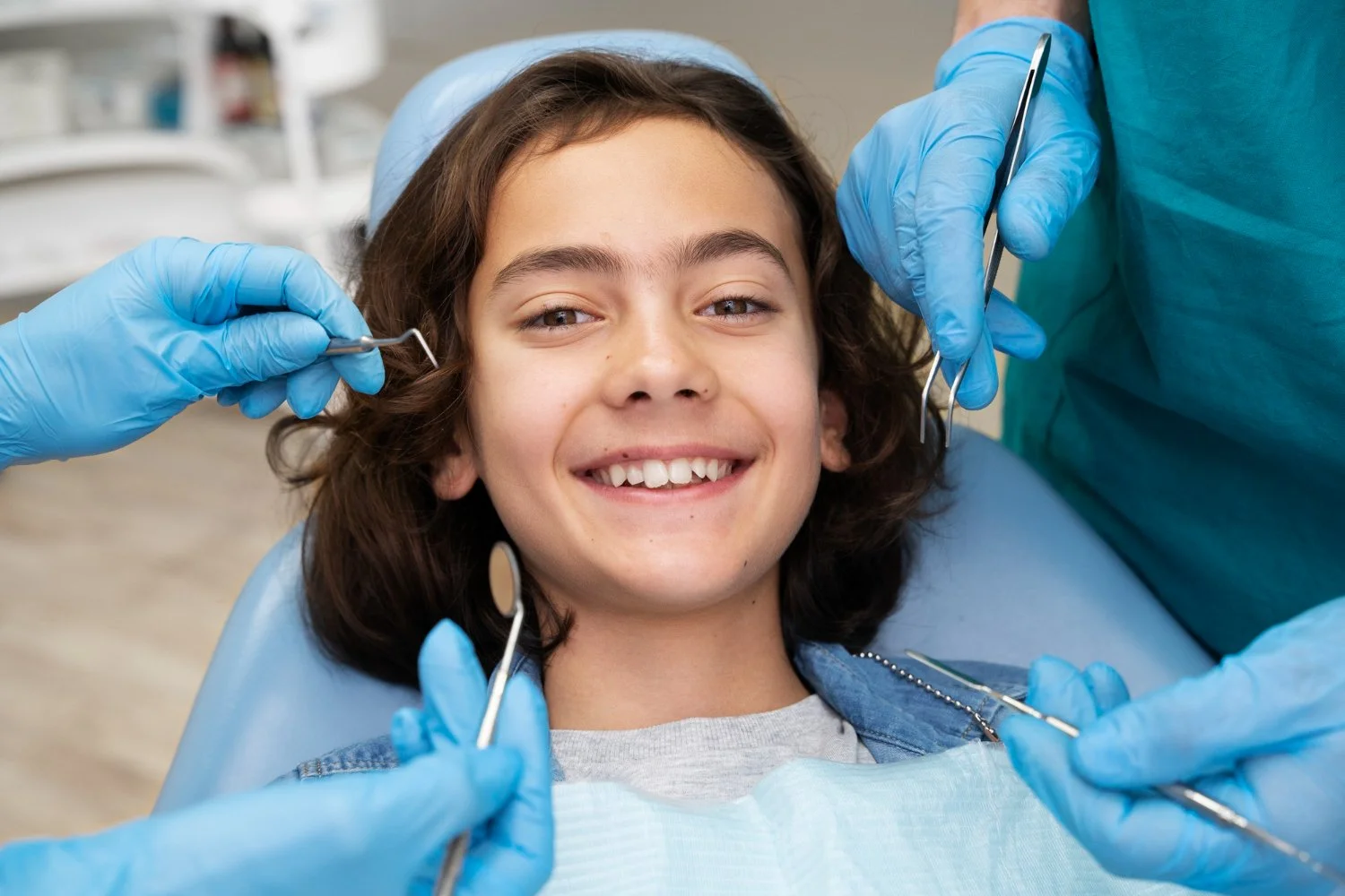 Young girl in dental chair smiling during dental examination with dentist wearing blue gloves holding dental tools.
