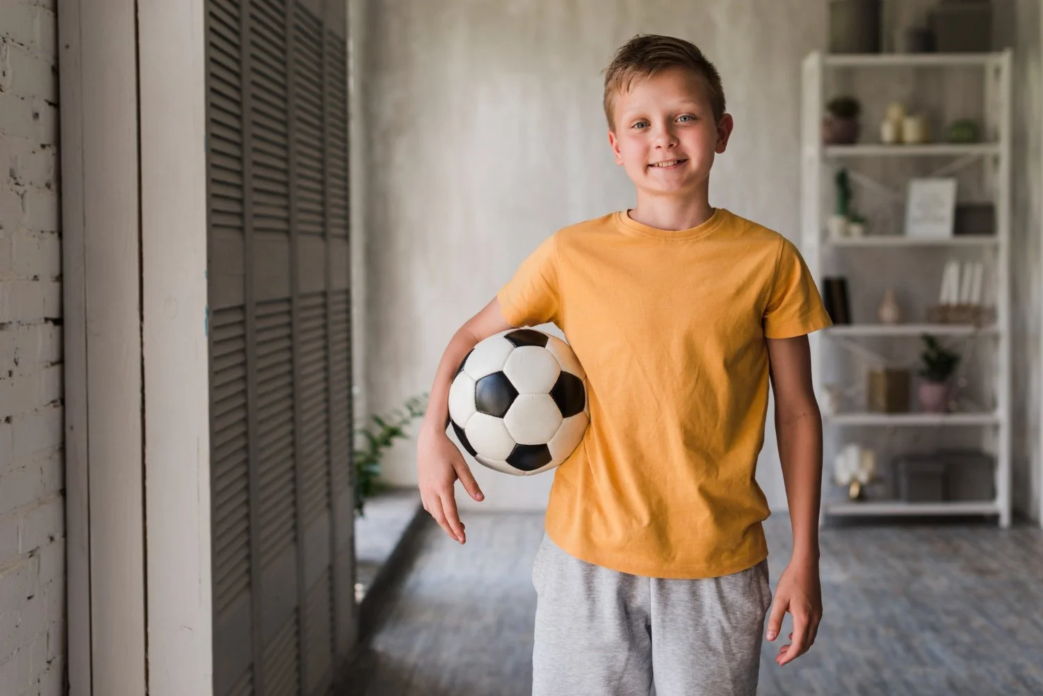 A young boy in a yellow t-shirt and gray sweatpants holding a soccer ball inside a room with white walls and a shelving unit in the background.