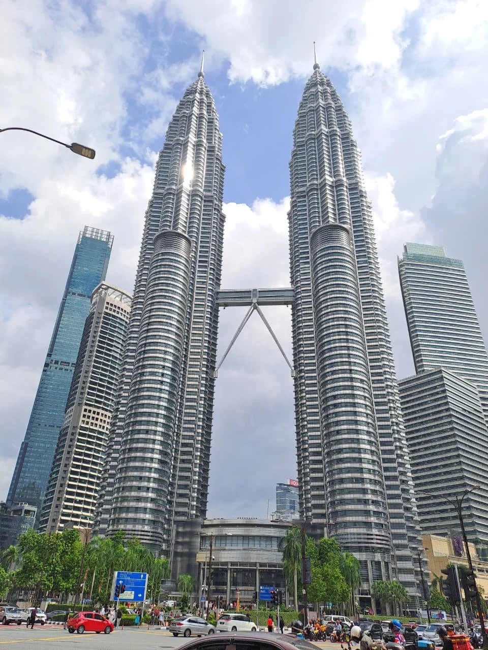 The Petronas Towers, a twin skyscraper complex in Kuala Lumpur, Malaysia, connected by a sky bridge at the top, set against a cloudy sky with surrounding modern buildings, cars, motorcycles, and greenery in the foreground.