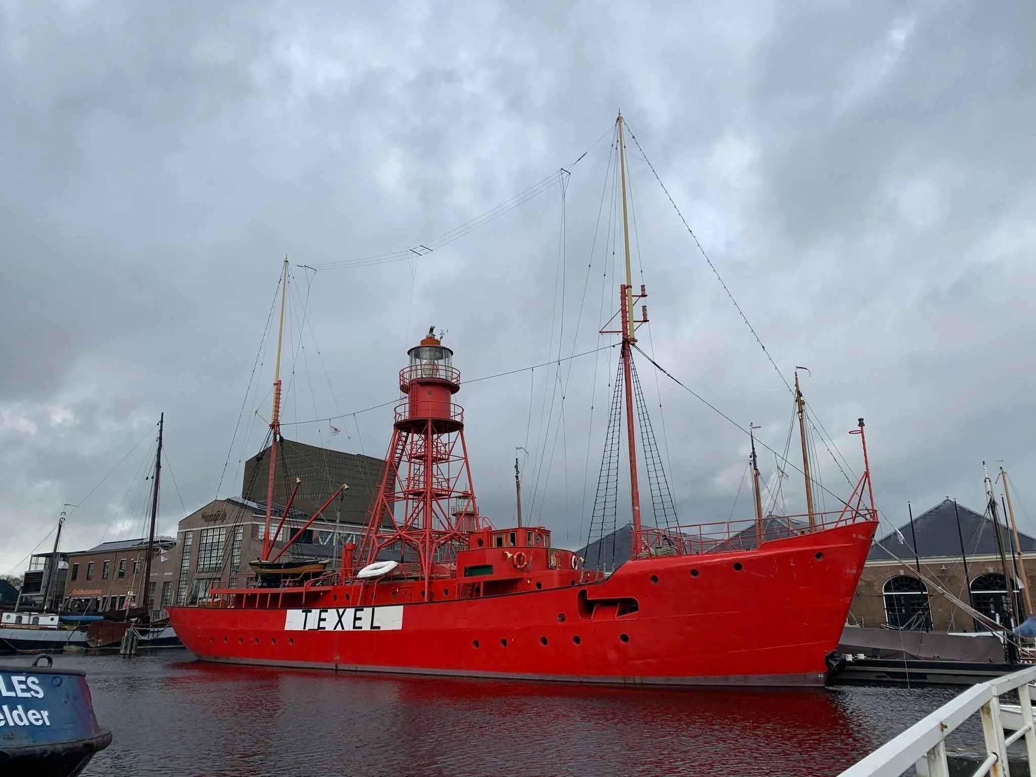 A large, red, historic lighthouse ship named TEXEL docked at a harbor, with gray overcast sky in the background.