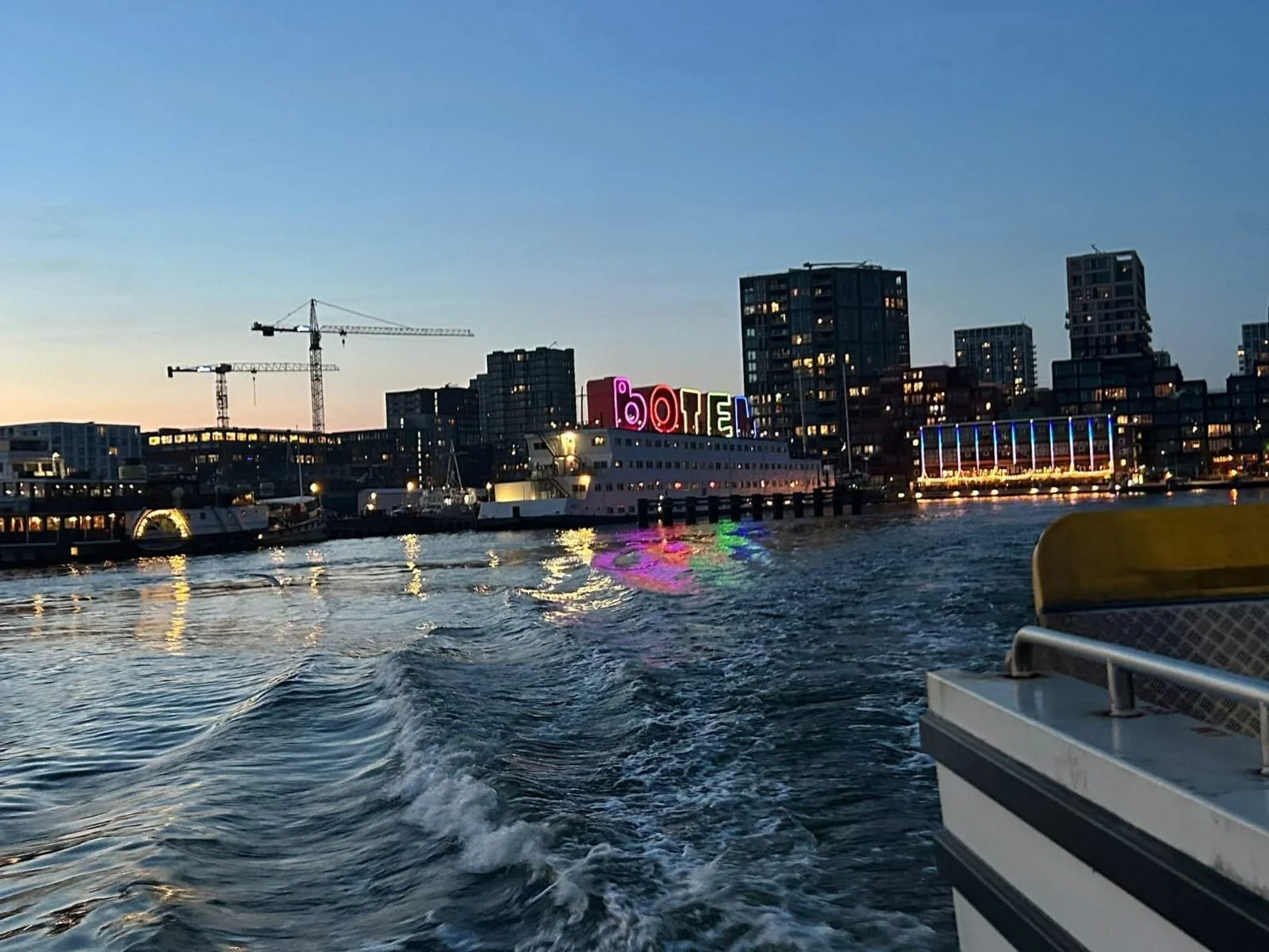 Waterway at dusk with boats and city buildings in the background. A cruise ship has a colorful sign on top spelling 'BOAT' and city lights reflect on the water.