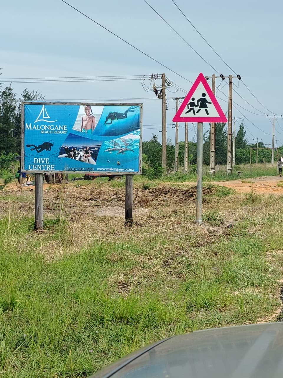 A roadside scene with a Malongane Beach Resort advertisement sign featuring images of diving, swimming, and a smiling boy. Next to it is a traffic sign indicating a pedestrian crossing. The background includes power lines and a dirt road surrounded b