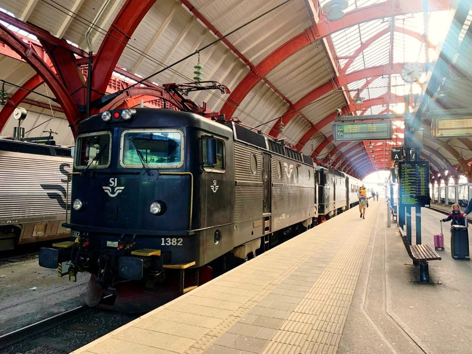 A black electric train sitting at a station platform with a few passengers and luggage, red arches in the station ceiling, digital departure screens, and a clock overhead.
