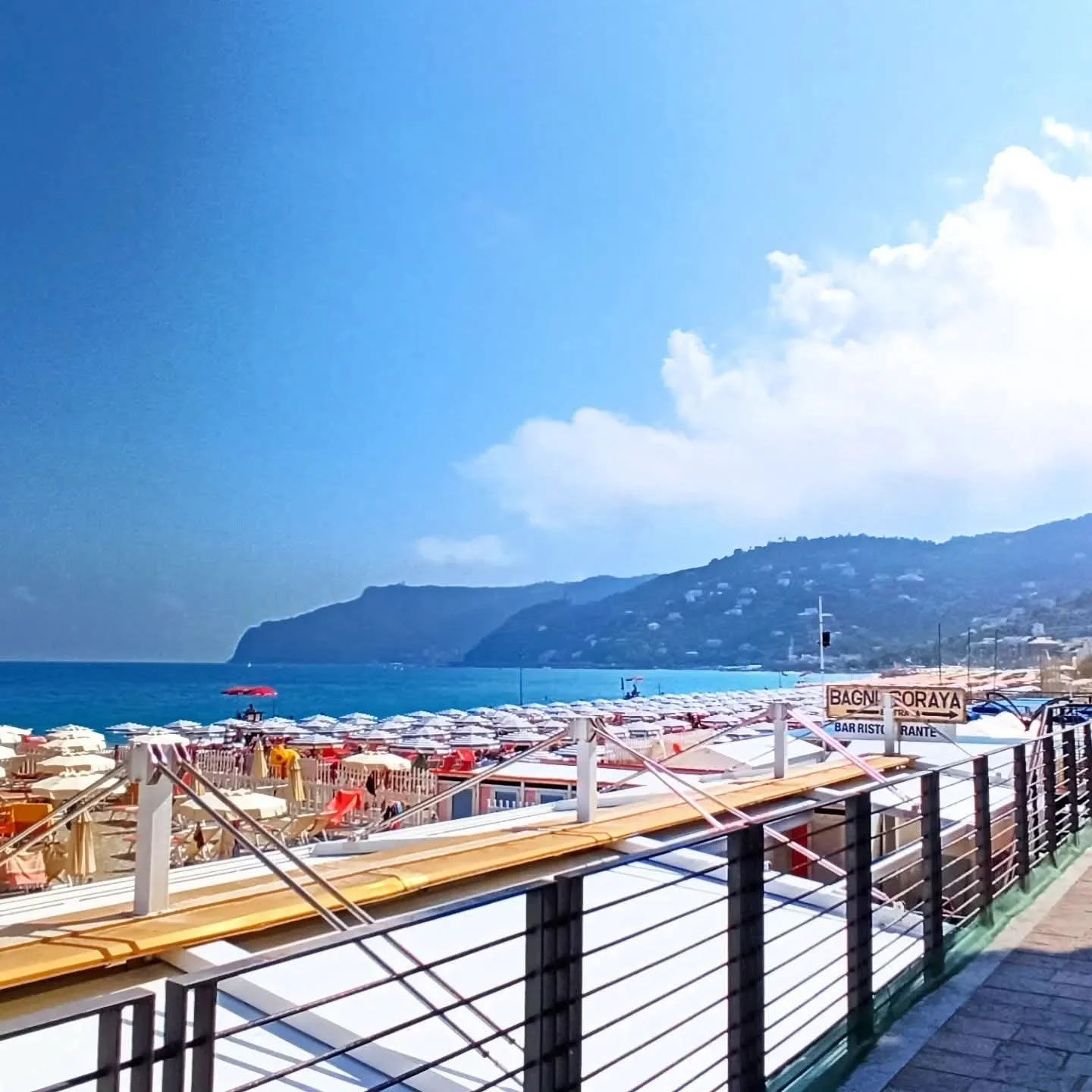 View of a beach with numerous umbrellas, umbrellas and lounge chairs, seen from a boardwalk with a railing. The sea and cliffs in the background under a partly cloudy sky.