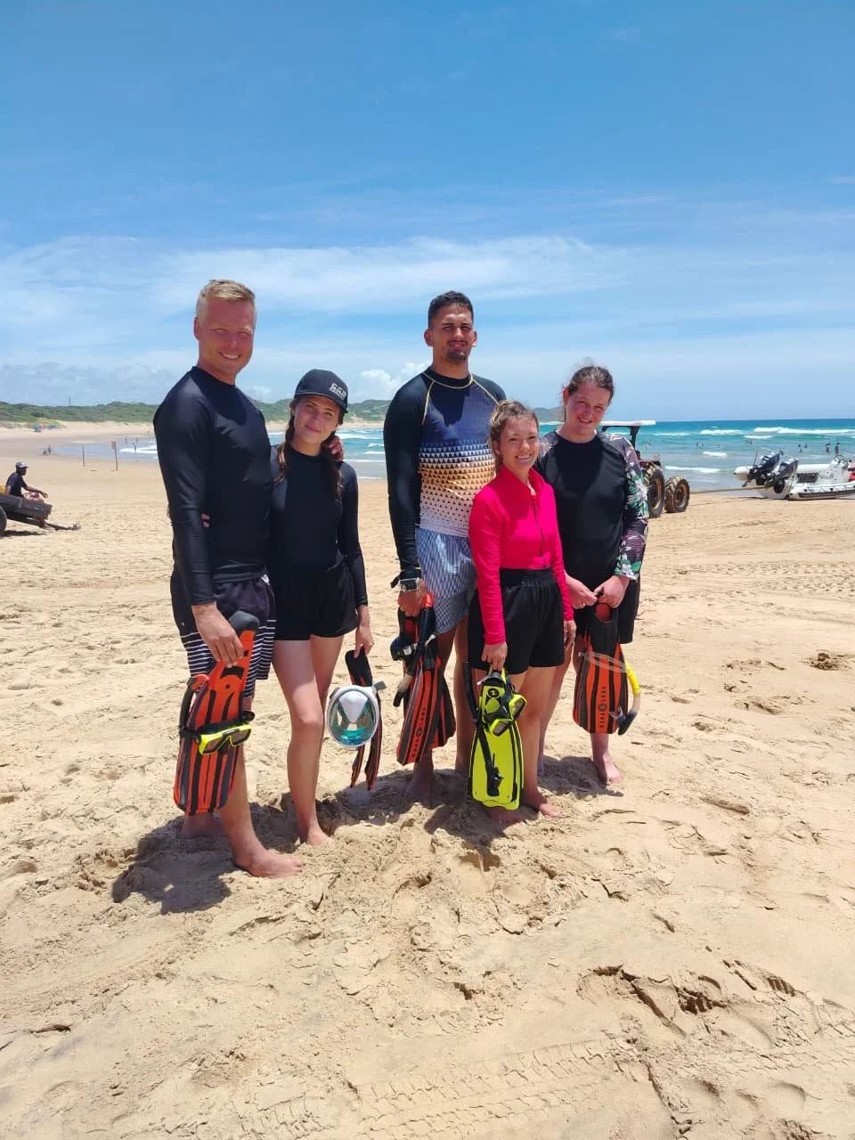 Group of five people standing on a sandy beach, wearing wetsuits and holding water sports equipment, near the ocean with blue sky and some boats in the background.