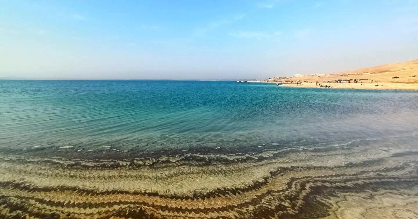 Beach with clear blue water and sandy shoreline, with land and sparse vegetation in the distance under a bright sky.
