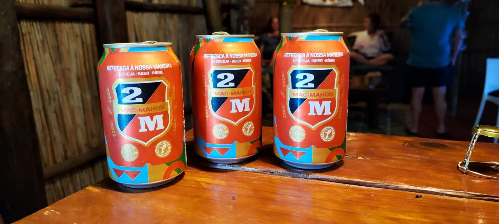 Three brightly colored cans of Mac Mahon beer on a wooden table in a rustic, indoor setting with a wooden wall in the background. Two people are sitting and standing in the background.