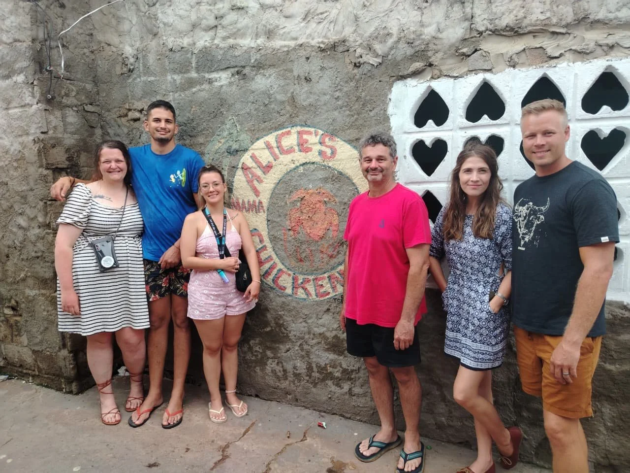 Group of seven people standing in front of a weathered wall with a painted sign that reads 'ALICE'S' and features a rooster graphic, some rockwork, and a wall with decorative cutouts behind them.