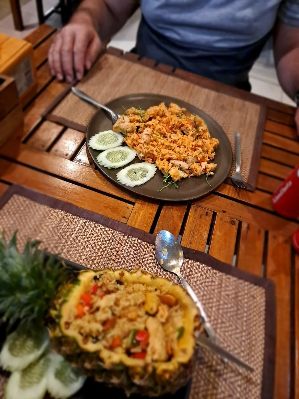 A person sitting at a wooden table with a cooked dish in front of them. The dish appears to be fried rice with chicken, garnished with cucumbers.