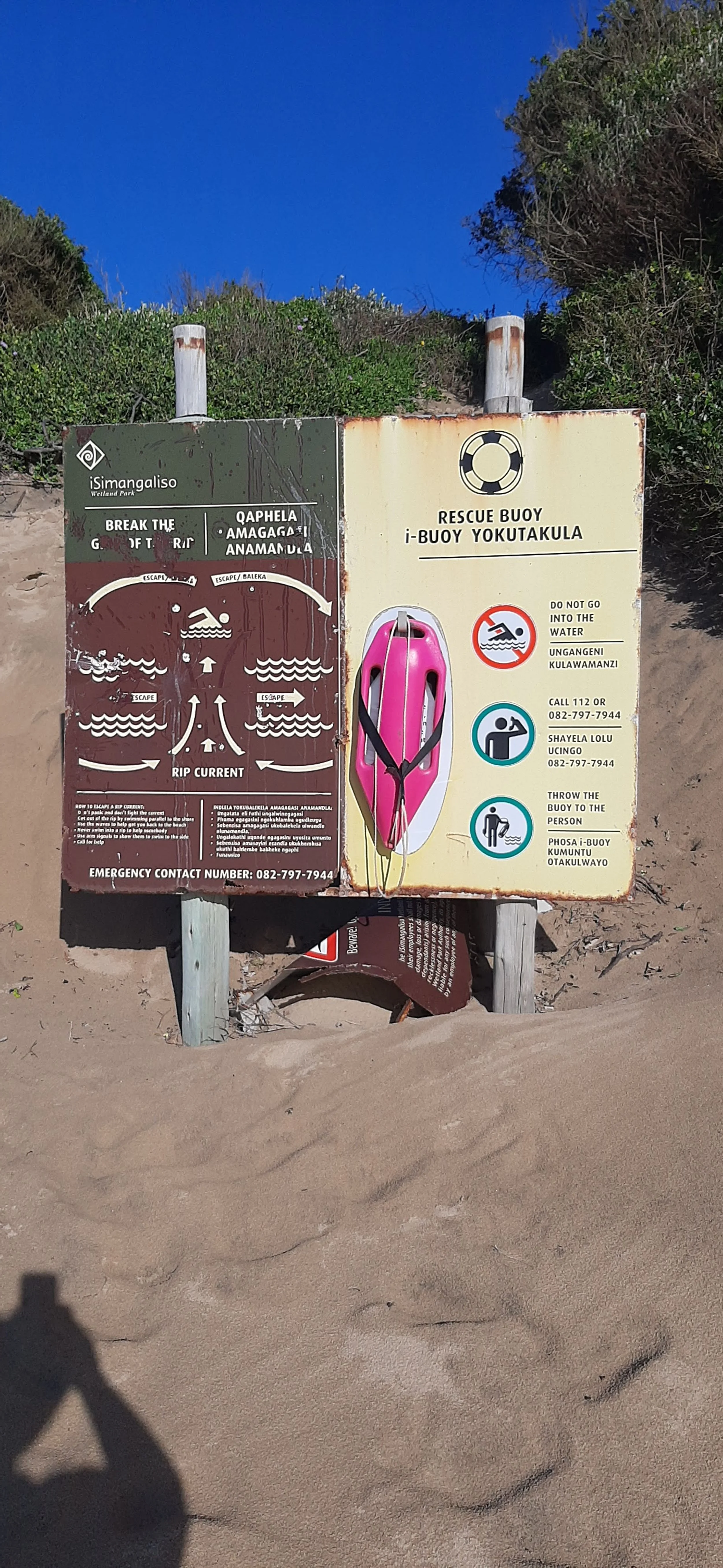Beach safety signs on the sand, including instructions for rip currents and rescue procedures, with a pink rescue buoy attached to the sign.