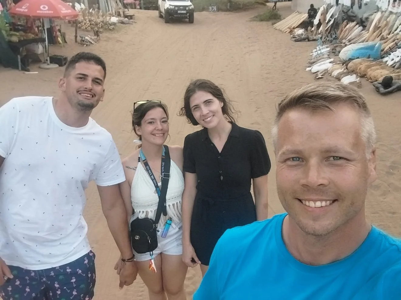 Four people smiling and posing for a selfie on a dirt path near market stalls with products, and a car in the background.