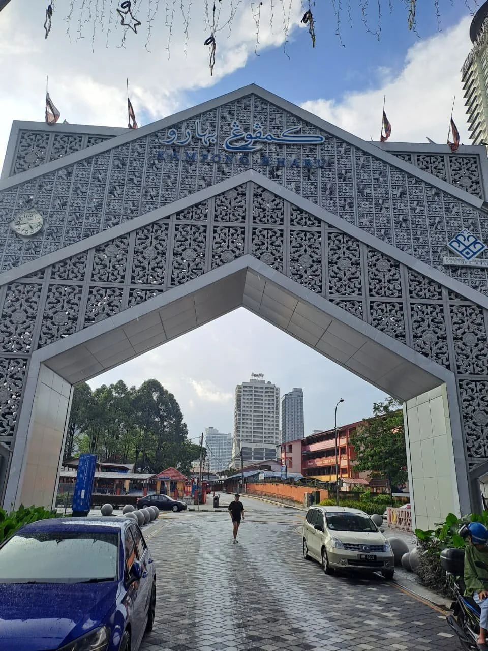 City street view through an ornate archway, with tall modern buildings in the background, parked cars and a person walking on a wet sidewalk.