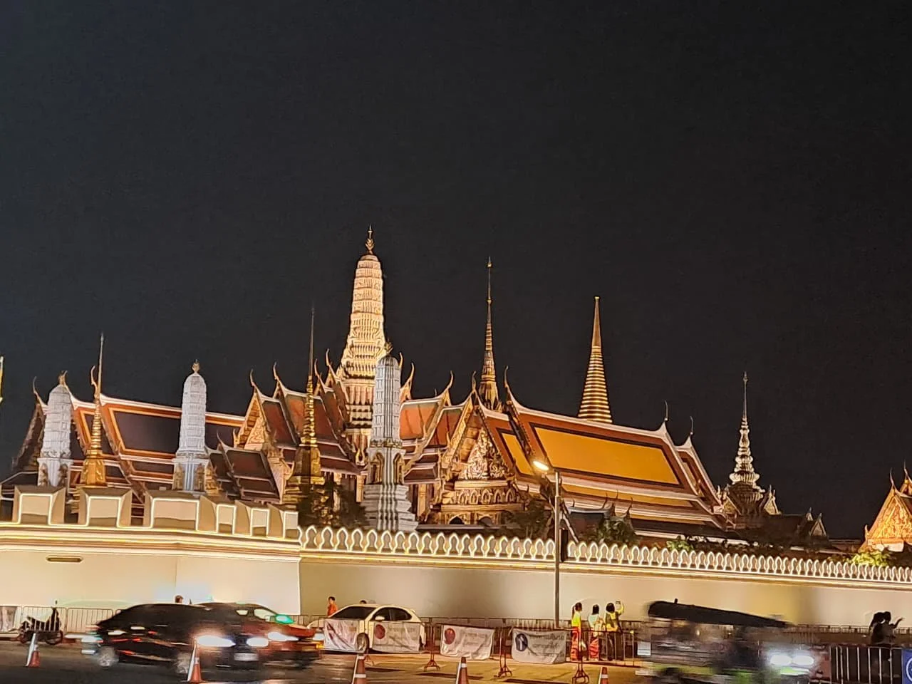 Night view of a traditional Thai temple with ornate rooftops and spires, illuminated behind a street with moving cars and pedestrians.