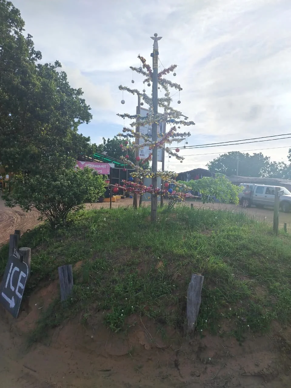 A decorated outdoor Christmas tree with ornaments and tinsel, situated on a small grassy hill next to an ICE sign, with a market in the background.