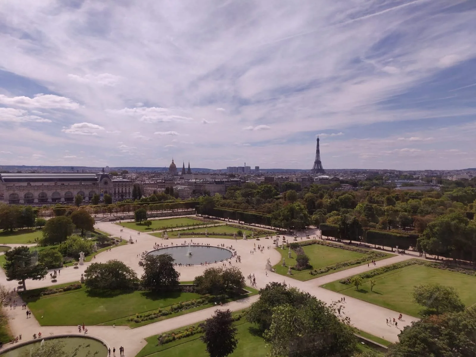 A panoramic view of Paris cityscape, including the Eiffel Tower, Sacré-Cœur Basilica, and a formal park with a fountain, pathways, trees, and visitors.