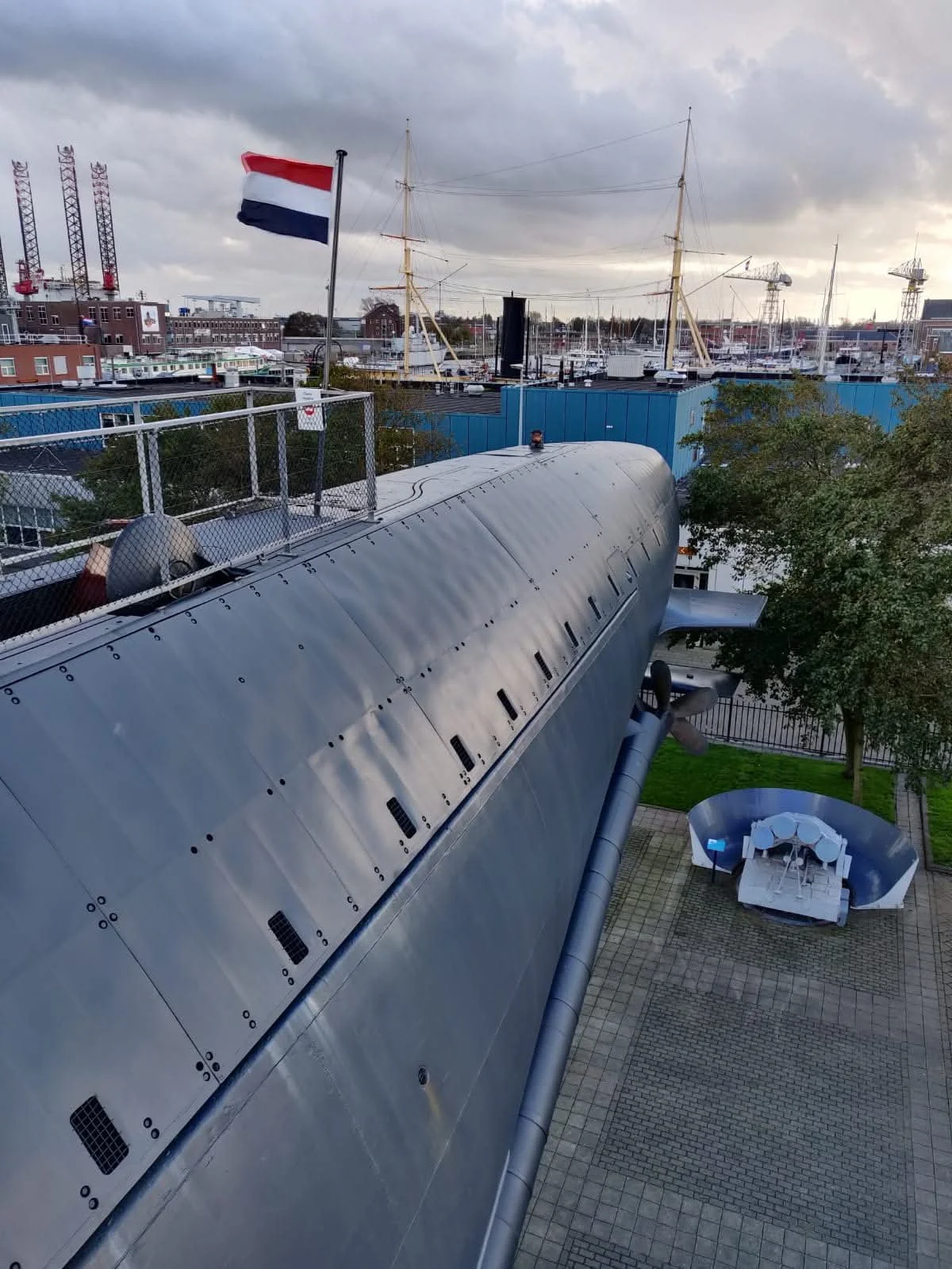 View of a large, metallic, curved structure from above, with a flagpole and Dutch flag, trees, a circle of chairs and tables, and a marina with boats and ships in the background under a cloudy sky.