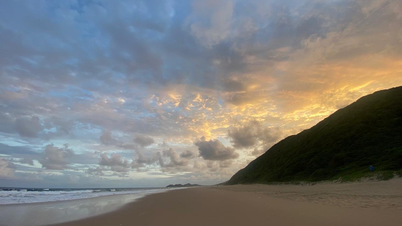 A sandy beach with waves crashing and a green hillside on the right under a partly cloudy sky at sunset.
