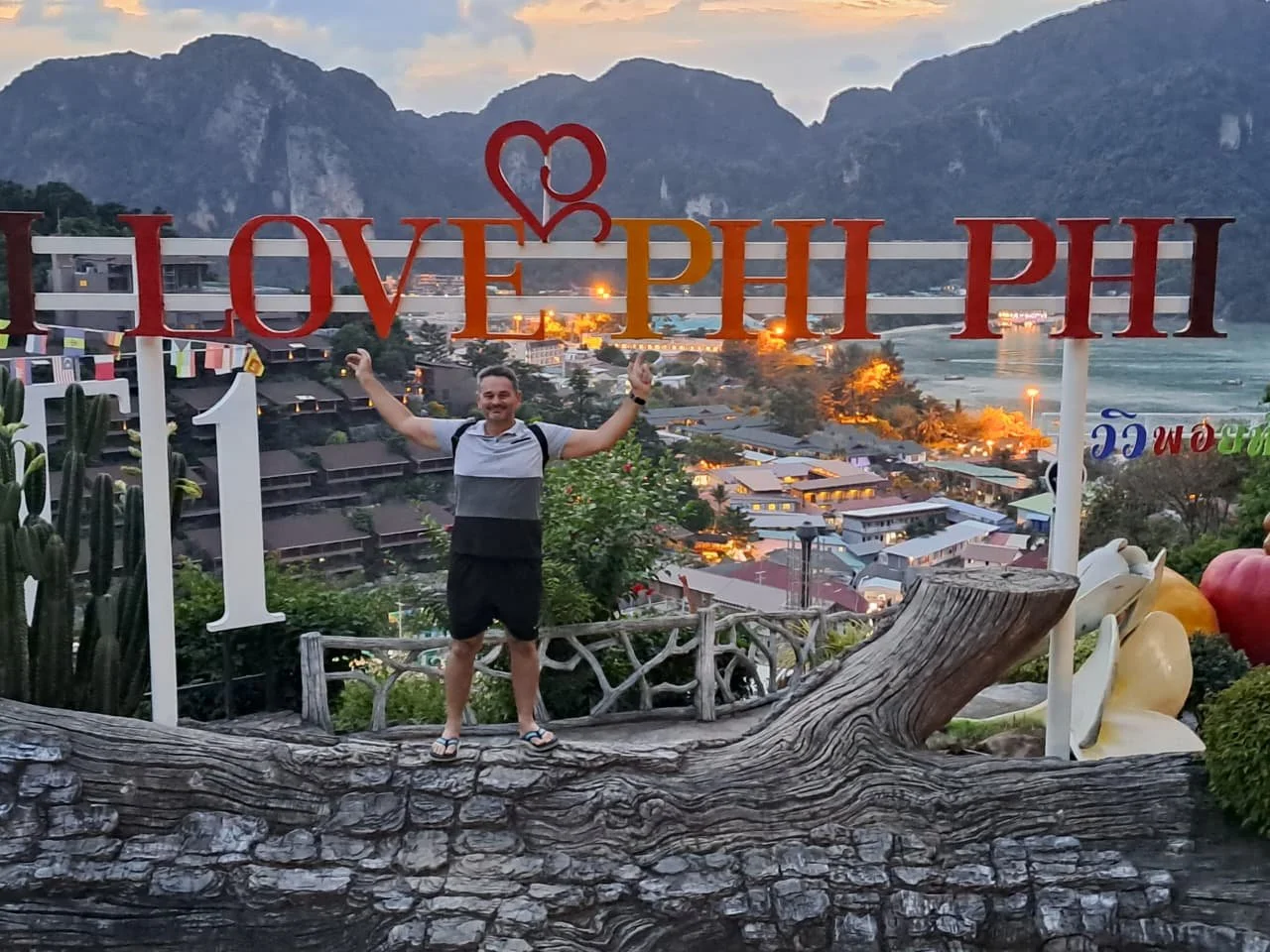 A man standing on a large tree trunk sculpture with their arms raised, smiling at the camera, in front of a scenic view of a town at sunset, with mountains in the background, and a glass sign reading "LOVE PHI PHI" with a heart-shaped design.