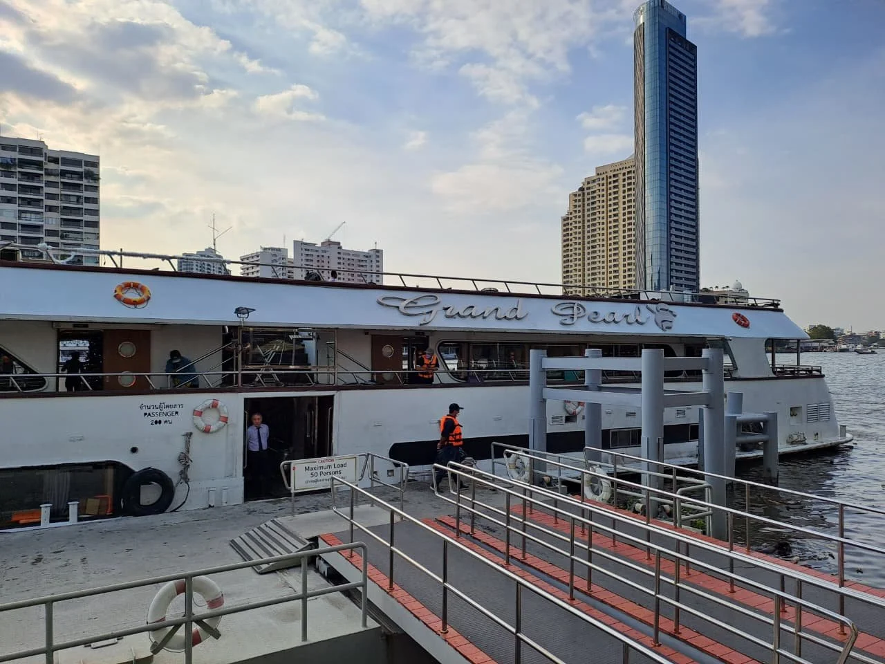 A large white passenger ferry named 'Grand Pearl' docked at a pier with city skyscrapers in the background, and staff members in orange uniforms near the entrance.