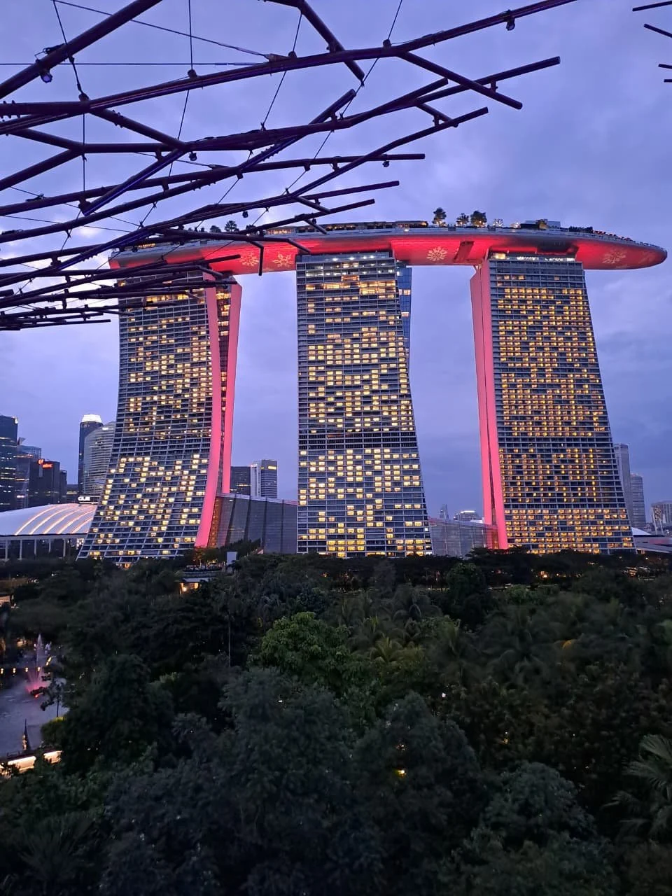 Night view of the Marina Bay Sands hotel in Singapore with three tall towers connected by a boat-shaped rooftop, illuminated with yellow lights and a purple-blue evening sky in the background.