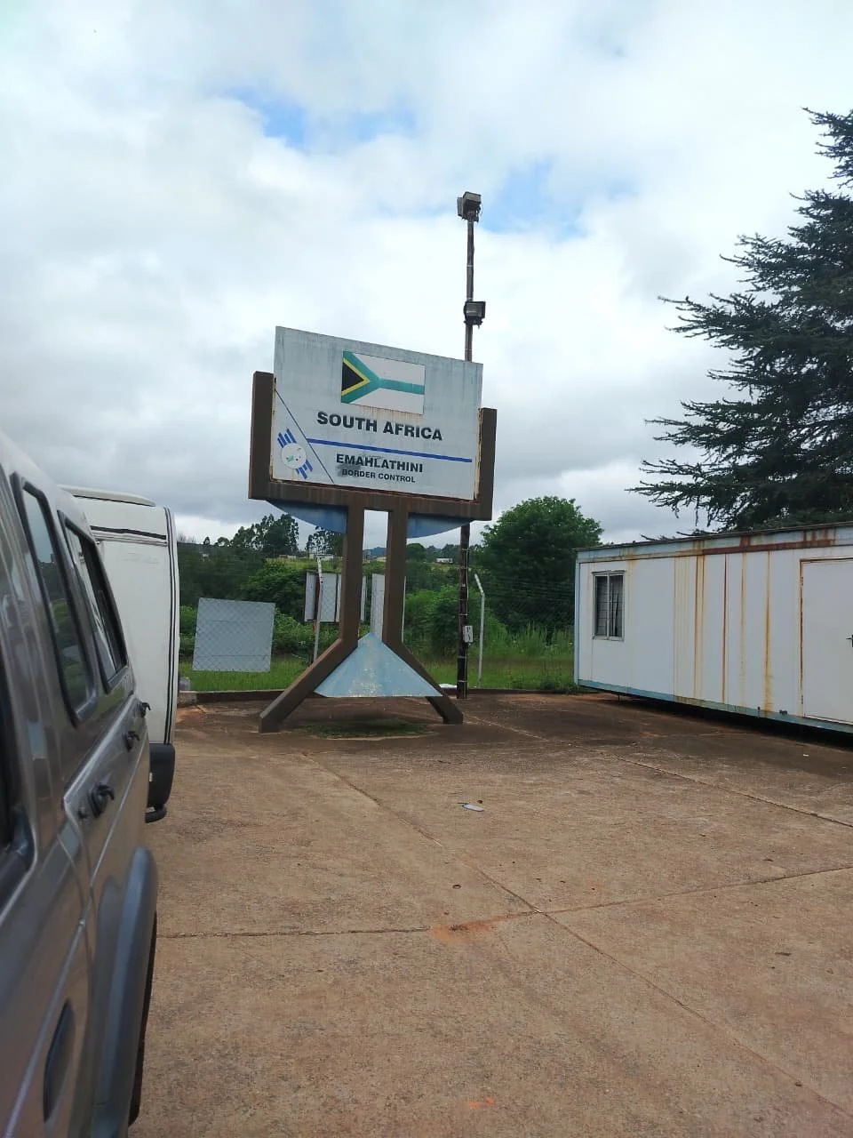 Sign indicating the border crossing between South Africa and Eswatini, with vehicles parked nearby and a background of trees and cloudy sky.