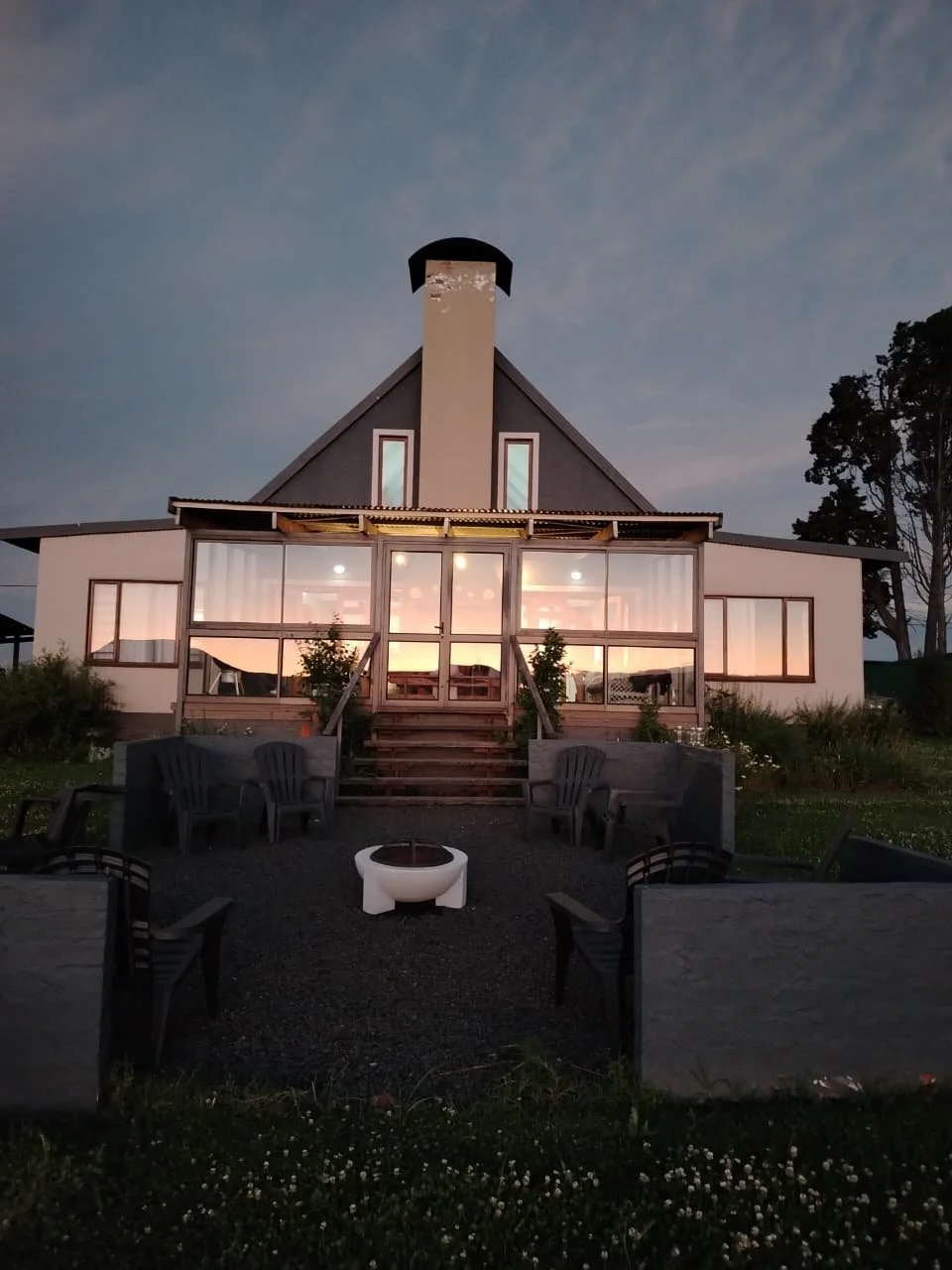 A modern house with large glass windows illuminated from inside, dark sky in the background, outdoor seating with chairs and a fire pit in the foreground.