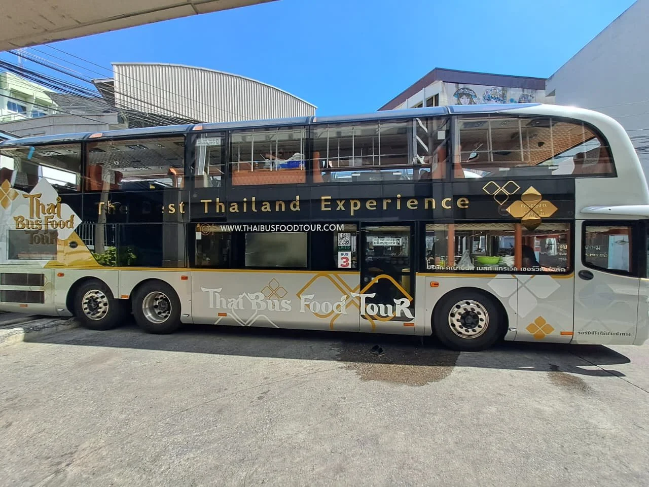 Double-decker bus advertising a Thai food tour with the text 'The Best Thailand Experience' and 'Thai Bus Food Tour' on its side, parked on the street with buildings and power lines in the background.