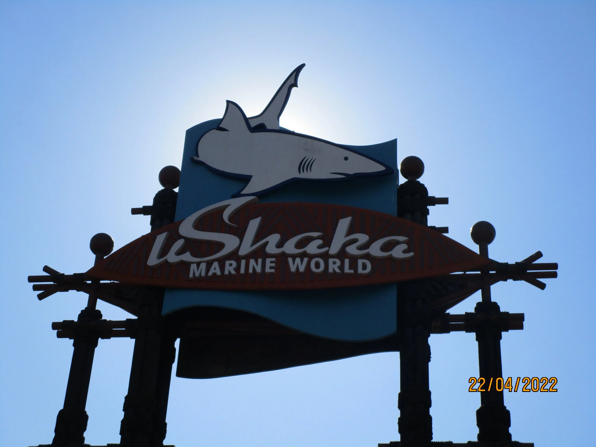 Sign for iShaka Marine World with a shark graphic, set against a bright blue sky.