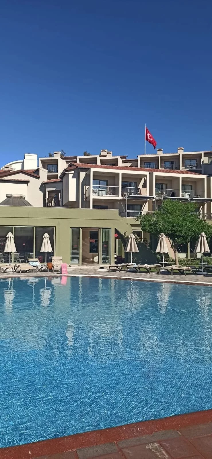 Swimming pool with lounge chairs and umbrellas in front of a multi-story building with balconies, a red flag with a white crescent moon and star, and a clear blue sky.