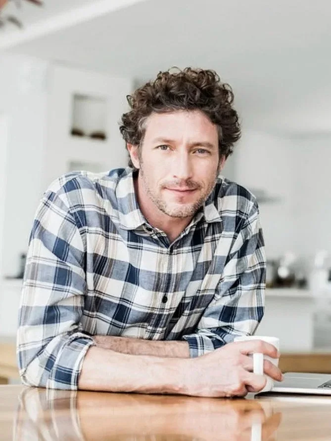 A man with curly hair and a beard, wearing a plaid shirt, sitting at a table in a bright, modern kitchen, holding a coffee mug.