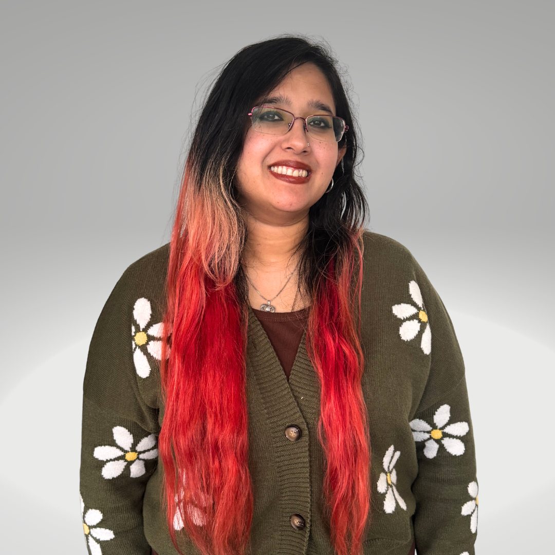 A woman with long, wavy hair dyed red at the ends, wearing glasses and a green cardigan with white daisies, smiling at the camera against a plain background.