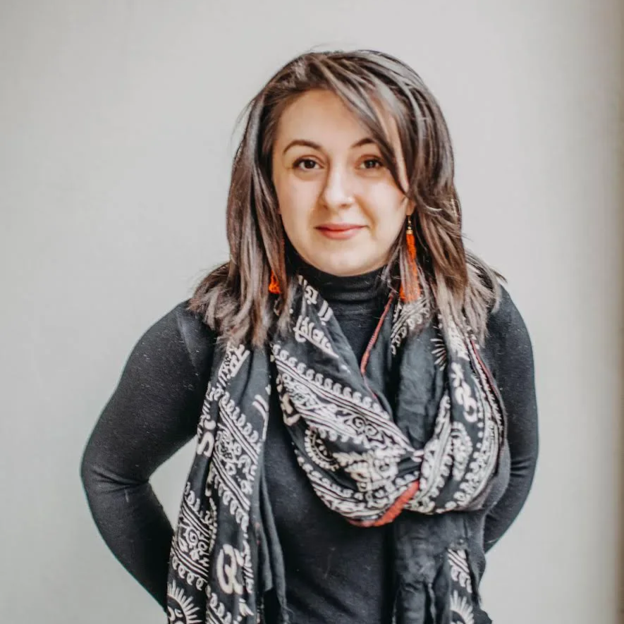 A woman with shoulder-length brown hair, wearing a black top, a black and white patterned scarf, and orange earrings, standing against a plain light gray background.