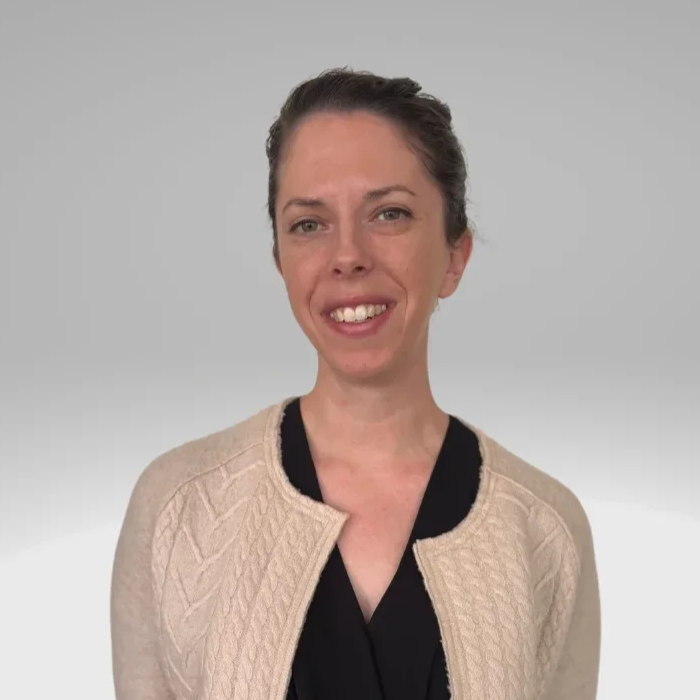 A woman with short dark hair, smiling gently, wearing a beige knitted jacket over a black top, standing against a plain light gray background.