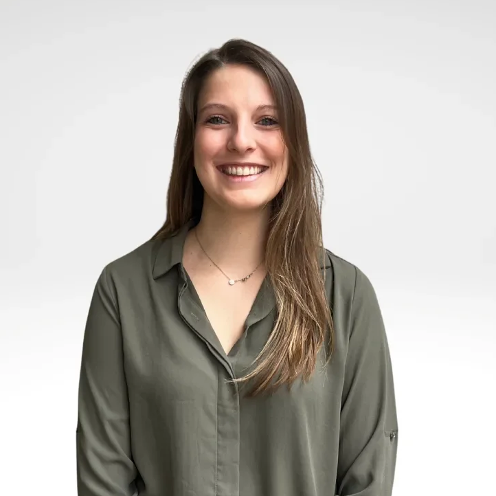 A young woman with long brown hair and a bright smile, wearing a green button-up shirt and a delicate necklace, standing against a plain white background.