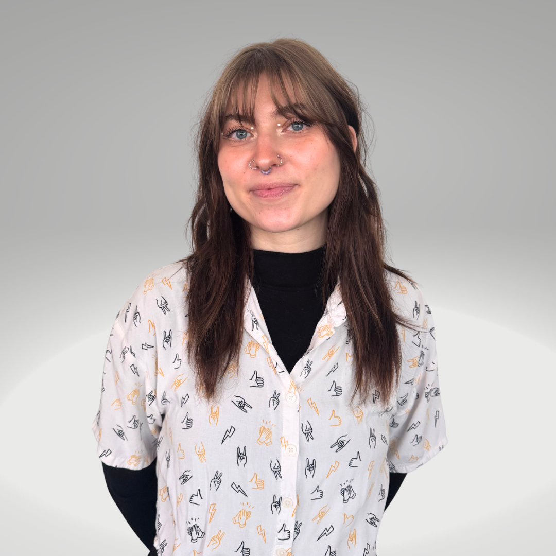 A woman with brown hair, blue eyes, and a septum piercing stands against a plain gray background, wearing a white button-up shirt with black and orange hand gesture icons over a black long-sleeve shirt.