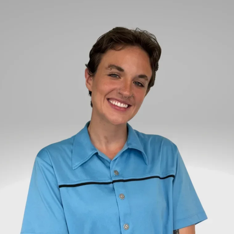 A young person with short dark hair wearing a blue button-up shirt, smiling, with a neutral gray background.