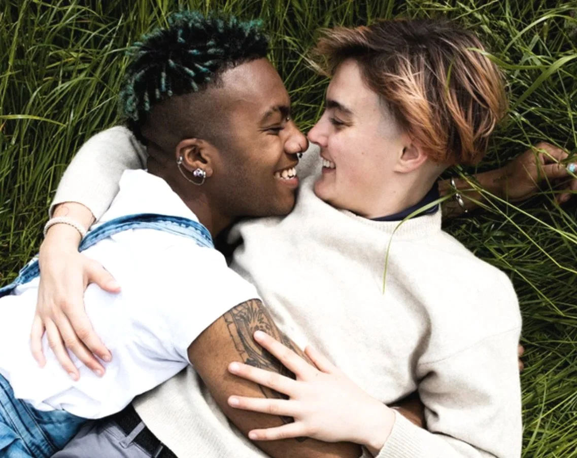 Two smiling men lying in grass, embracing and touching noses.