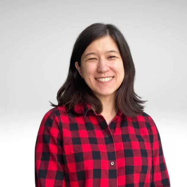 A smiling woman with shoulder-length dark hair wearing a red and black plaid shirt against a plain light gray background.